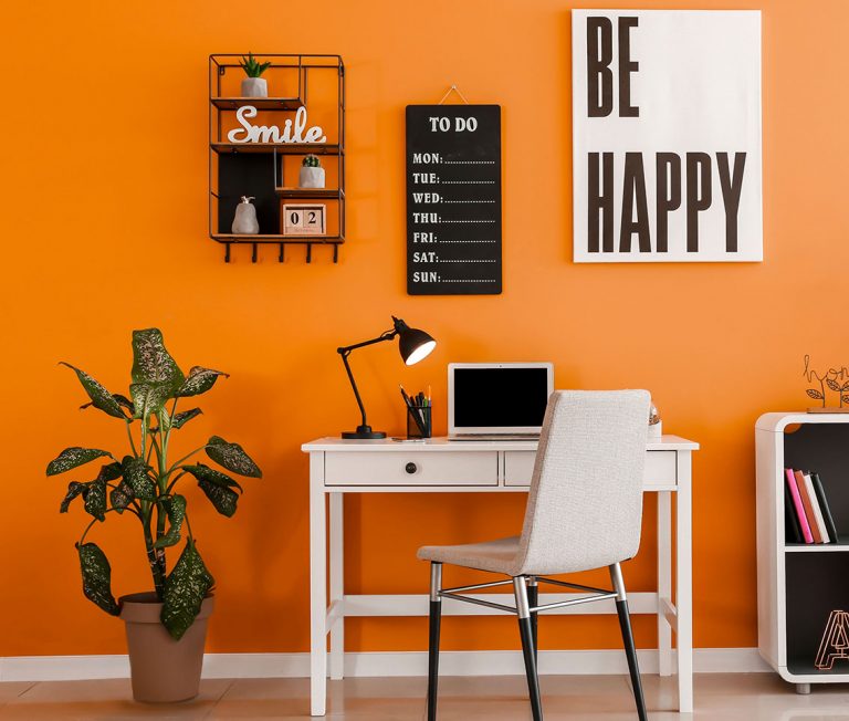 Arbeitsplatz mit orangener Wand, weißem Schreibtisch, Laptop, Pflanzen und Bildern