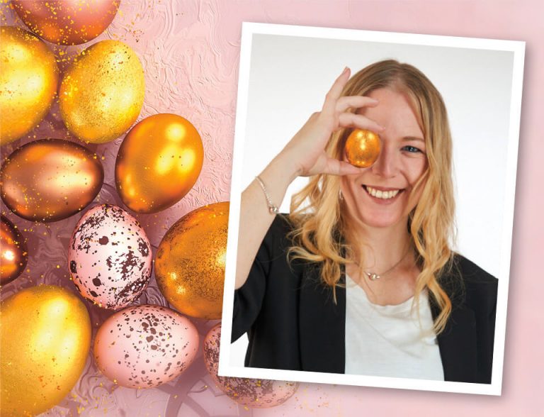 Elly lachend mit goldenem Ei vor dem Auge und im Hintergrund rosa und goldene Eier